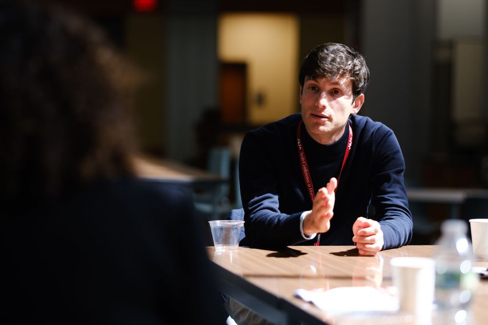 Young man in navy sweater engages in focused conversation at a conference table discussion.