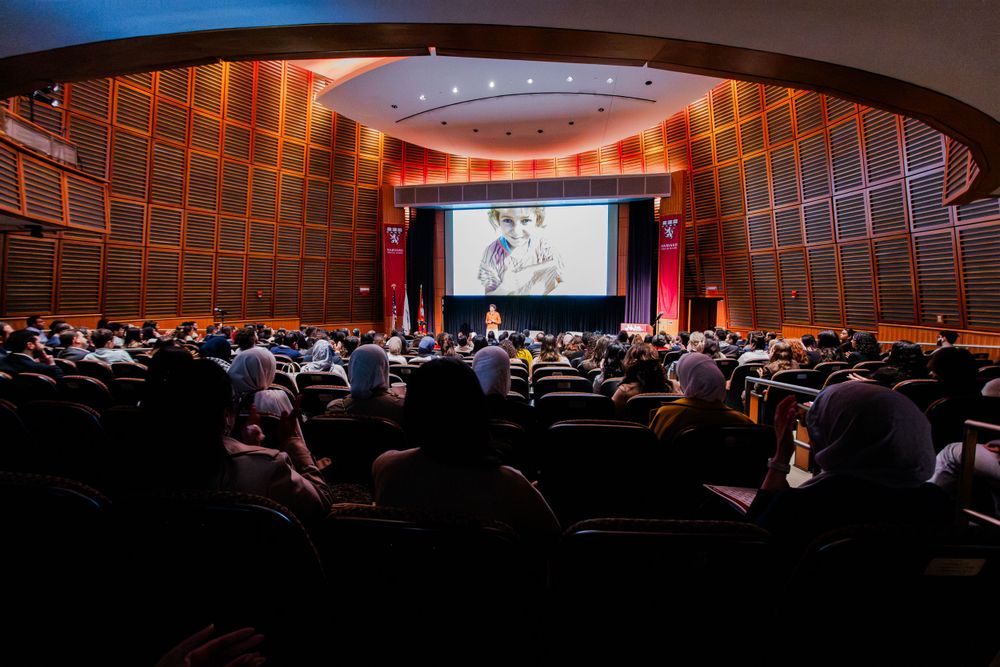 Wide-angle view of a packed auditorium audience watching a presentation on a large screen.