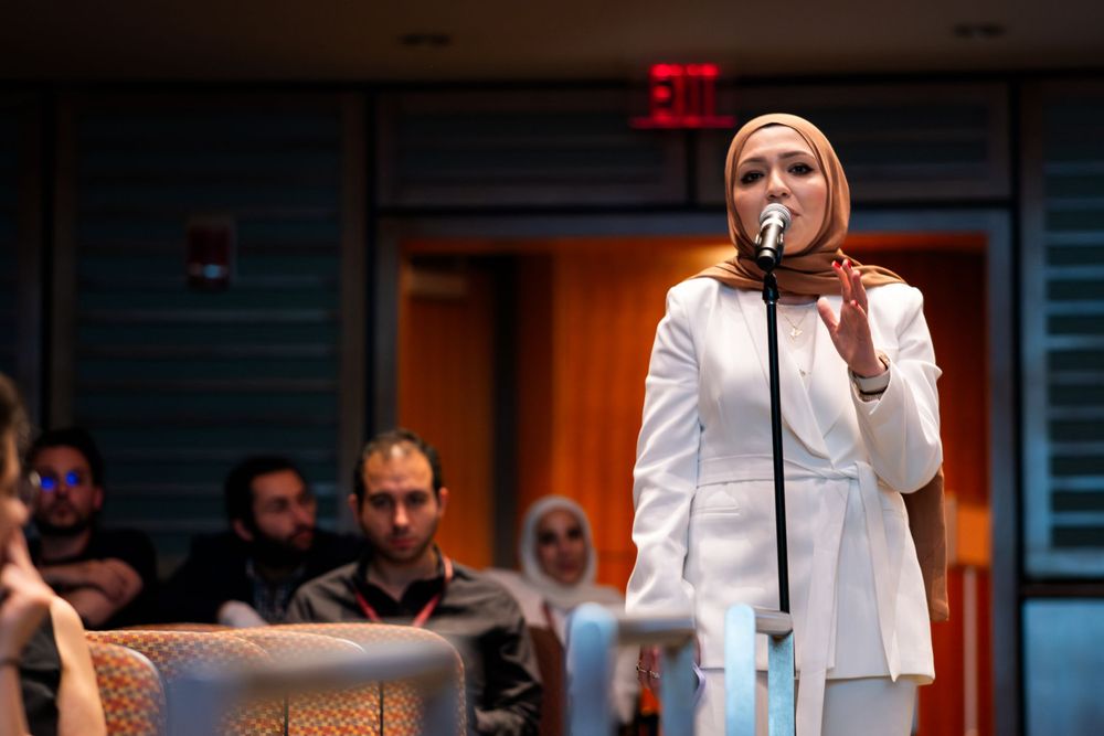 Woman in white hijab and suit speaks into a microphone at a conference presentation.