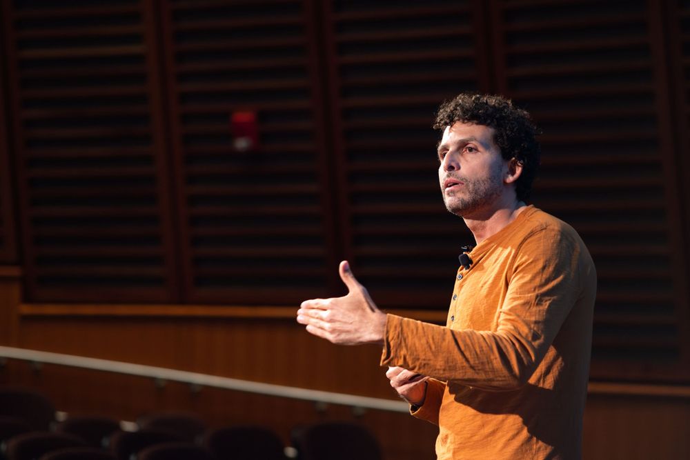 Man in orange shirt gestures while delivering a passionate speech on an auditorium stage.