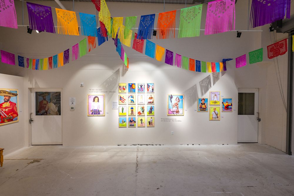 Art gallery exhibition space with colorful paper banners hanging from ceiling and artwork displayed on white walls.
