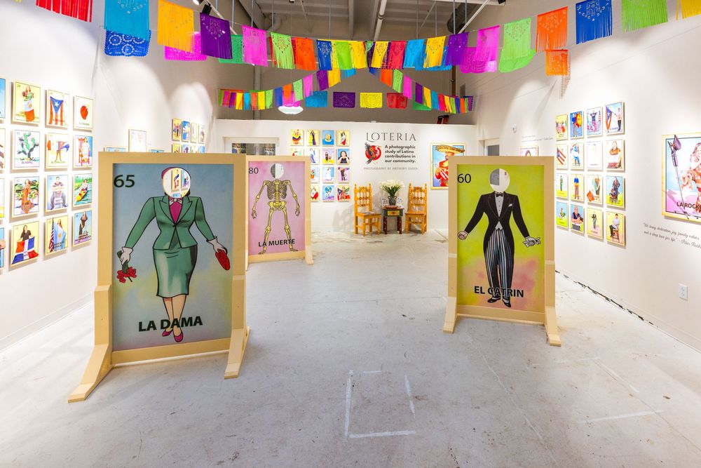 Colorful art gallery exhibition displays children's drawings and artwork on white walls with festive papel picado banners above.