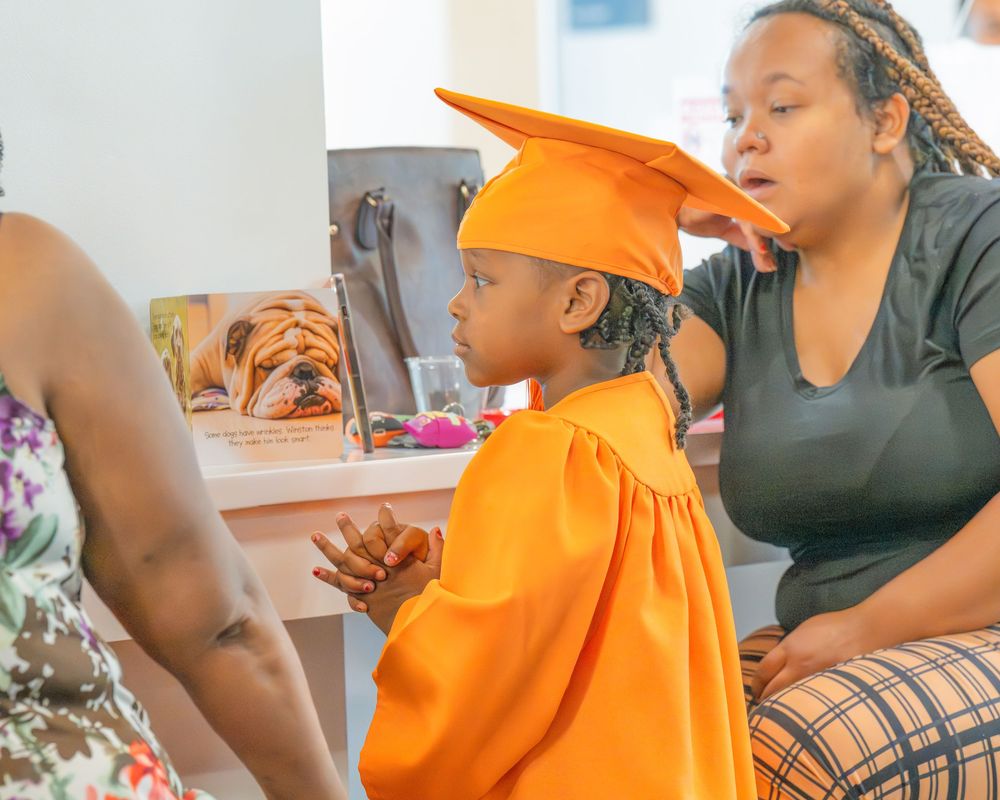 A child wearing a bright orange graduation cap and gown sits with others during a celebration ceremony.