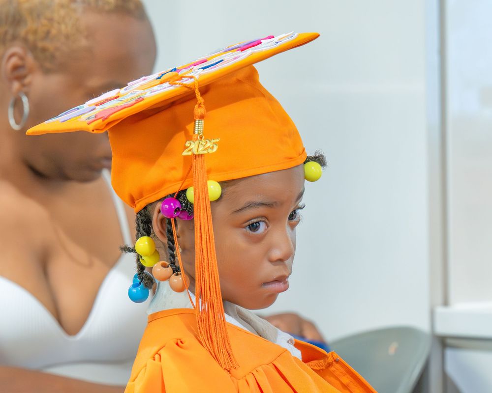 A graduate wearing a bright orange cap and gown looks thoughtfully to the side during commencement ceremonies.