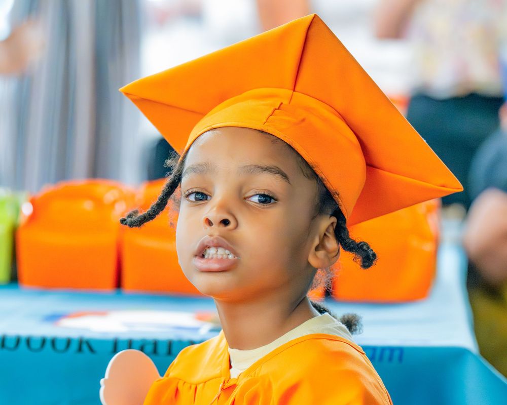 Graduate enjoying commencement ceremony while wearing bright orange cap and gown with a joyful expression.