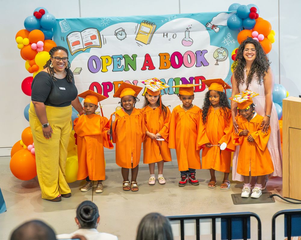 Children in orange graduation gowns stand in front of colorful Open A Book backdrop during a celebration ceremony.