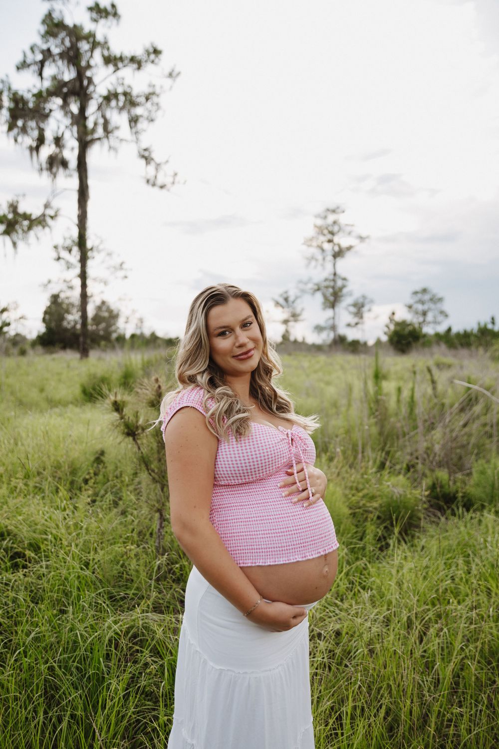 Maternity photo in a natural field setting with tall grass and trees shows an expectant mother in pink and white attire.