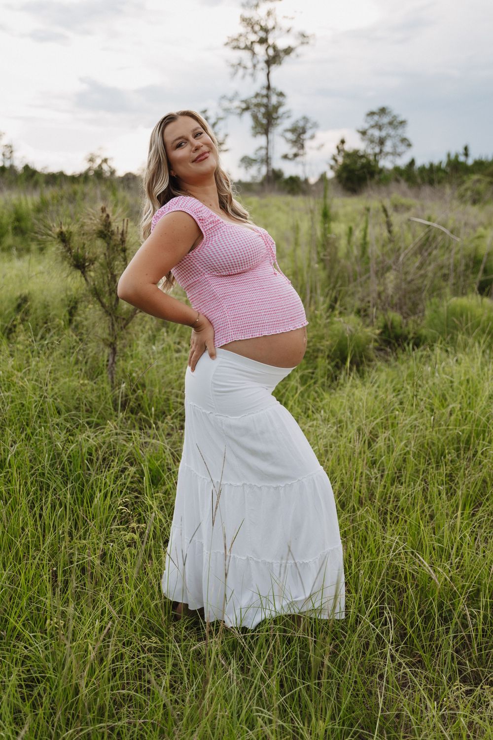 Woman in pink crop top and white skirt poses gracefully in tall grass field on sunny day.