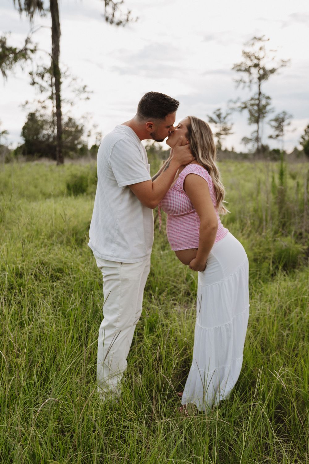 A romantic couple sharing an intimate moment in a grassy field at sunset, wearing white and pink attire.