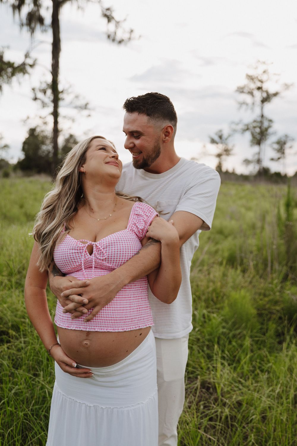 A romantic couple in pink and white outfits embraces in a green meadow during an outdoor photoshoot.