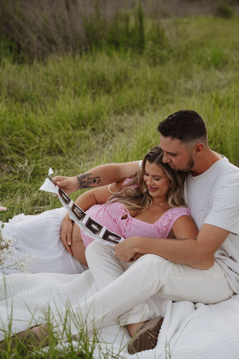 A couple shares an intimate moment sitting on a blanket outdoors, holding ultrasound photos while embracing.