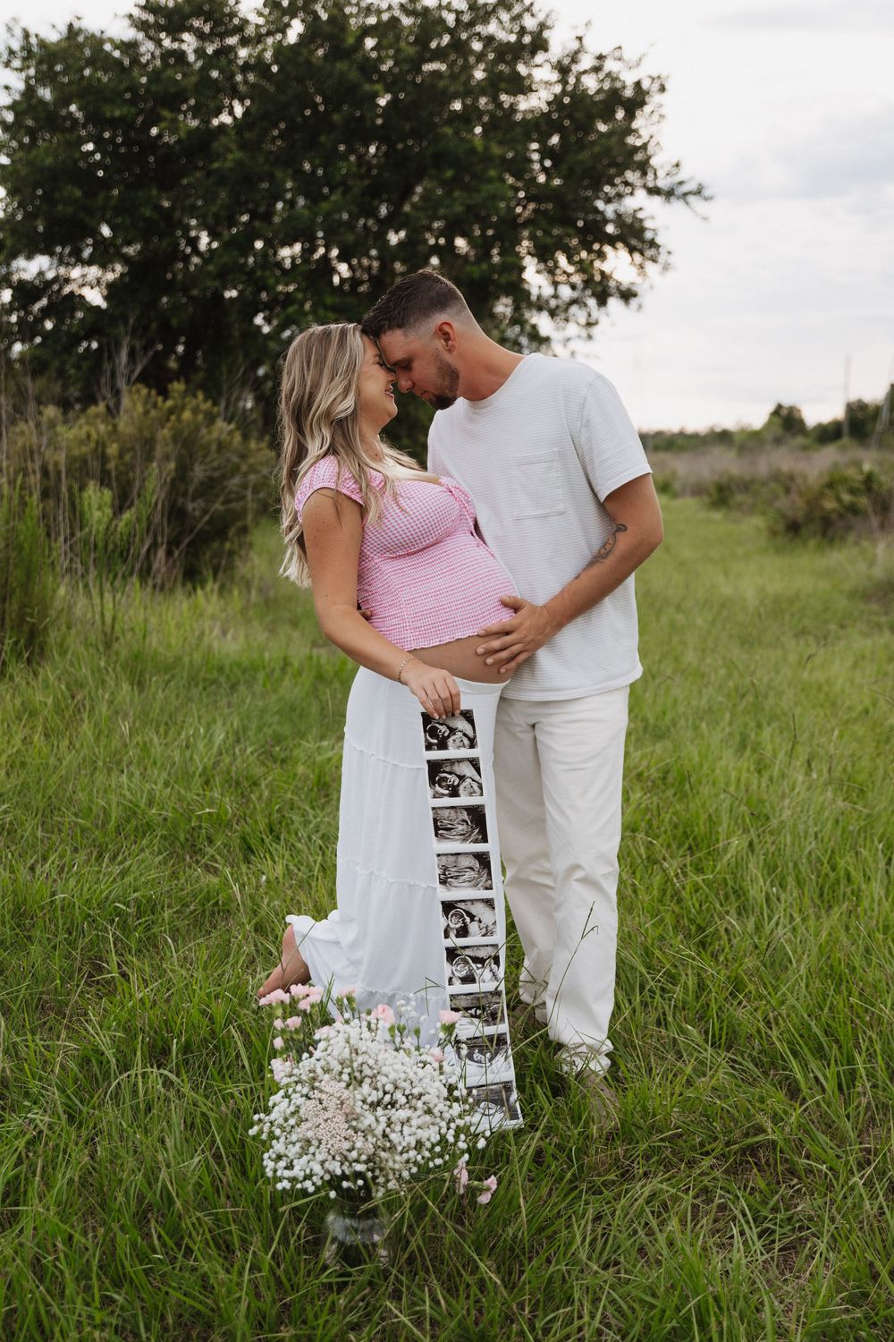 Couple sharing intimate moment in grassy field with ultrasound photos and baby's breath flowers for pregnancy announcement.