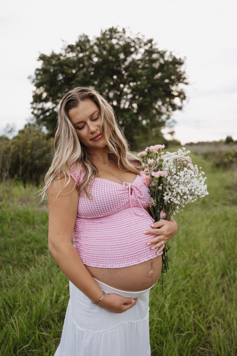 Woman in pink gingham crop top and white skirt holds wildflowers in outdoor field during golden hour.