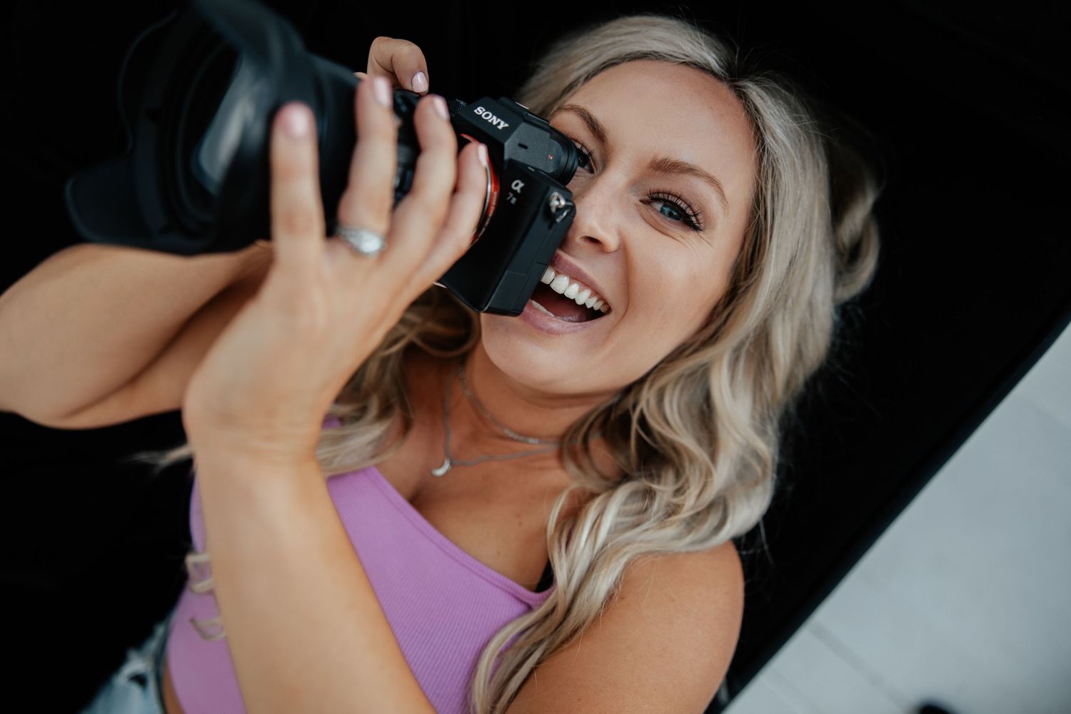 A photographer in purple holds a professional camera with enthusiasm against a dark background.