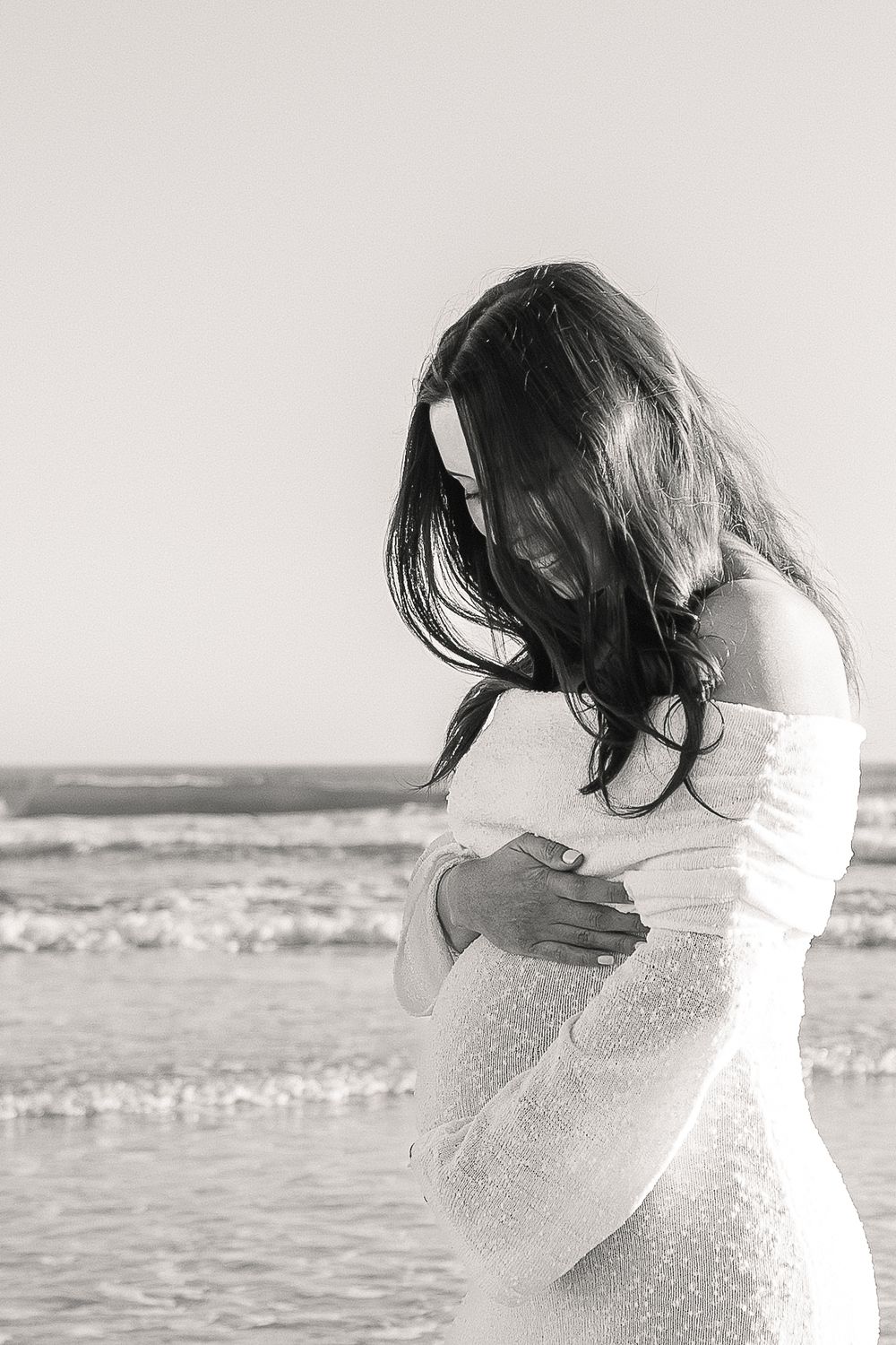 Black and white artistic photo of someone in a white dress standing by the ocean waves with windswept hair.