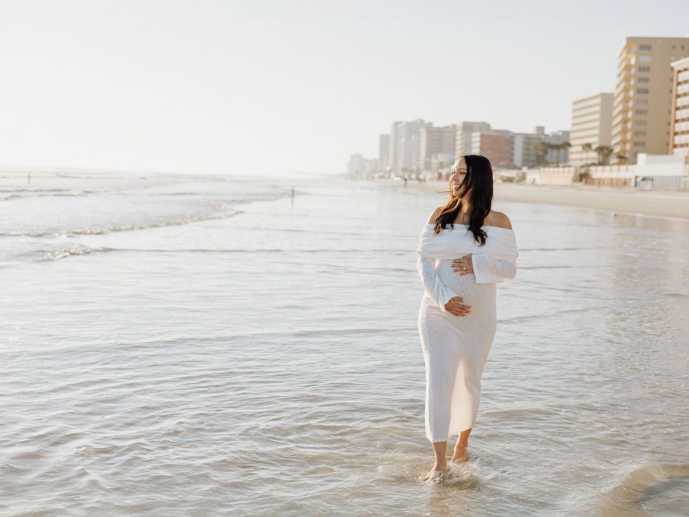A person in a white dress walks along a serene beach shoreline during sunset while waves gently roll in.