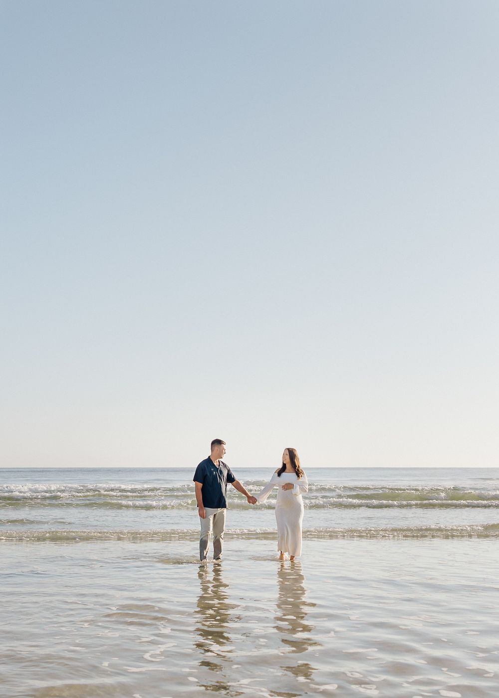 A couple walks together in shallow ocean waves at sunset holding hands on a serene beach.