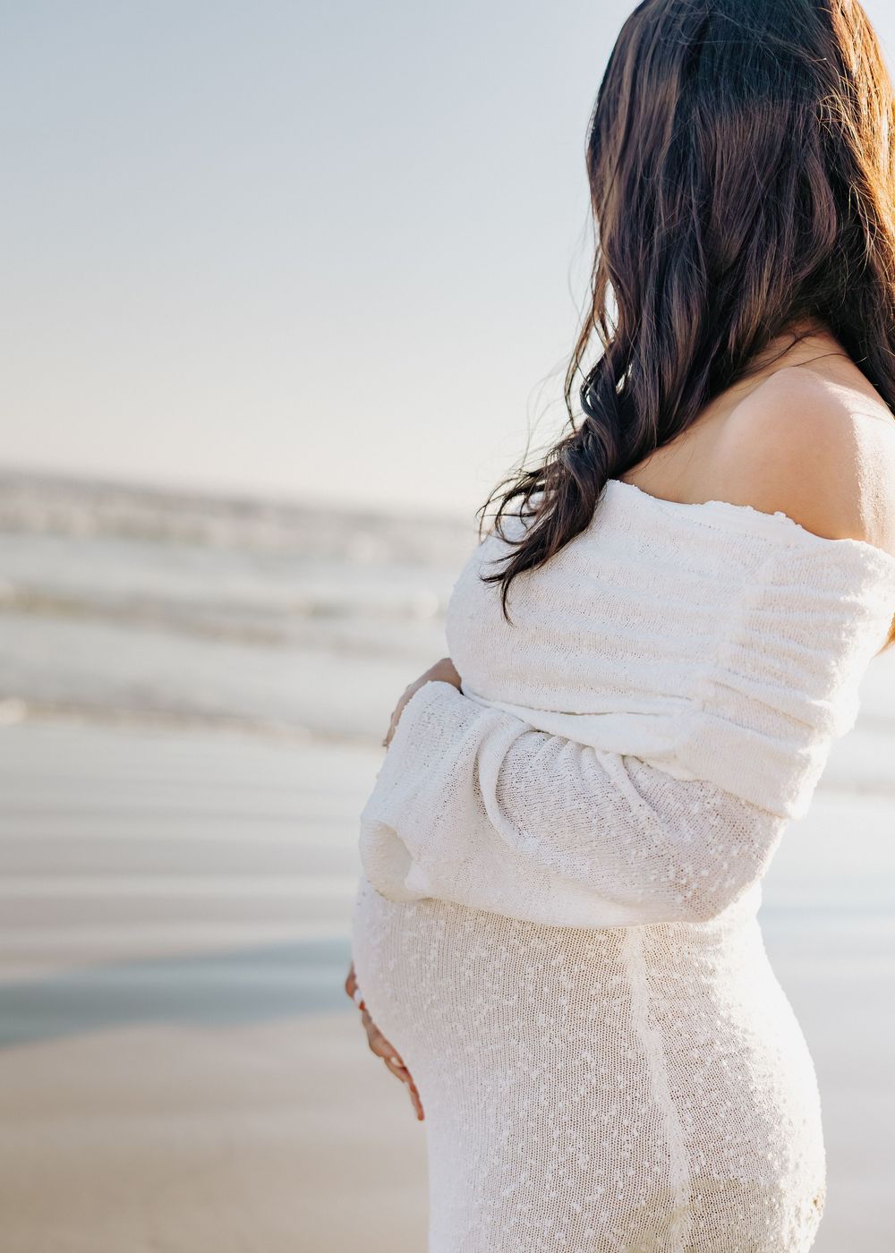 Side view of a pregnant figure in a white off-shoulder dress standing by the ocean at sunset.