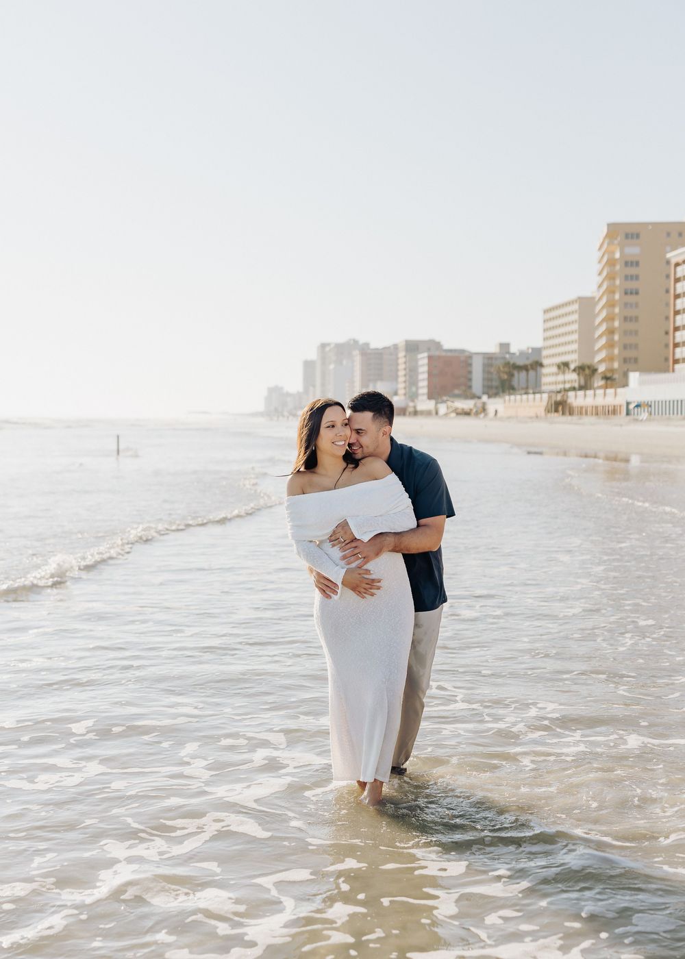 A couple embraces while standing in shallow ocean waves along a beachfront with high-rise buildings in the background.
