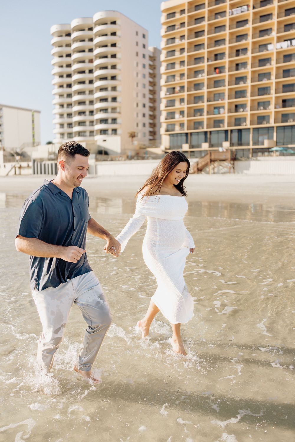 A couple plays together on a sandy beach with high-rise condominiums in the background during a sunny day.