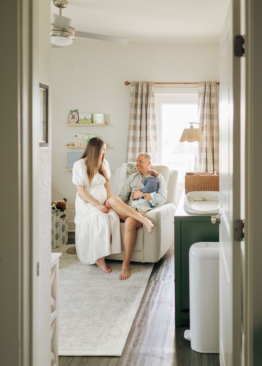 A woman in a white dress sits beside an older man holding a newborn in a cozy, softly lit nursery room.