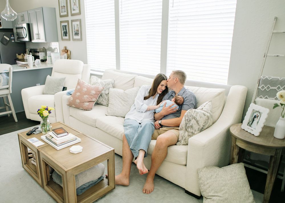 A couple sits on a white sofa, lovingly gazing at their newborn baby in a bright, cozy living room.