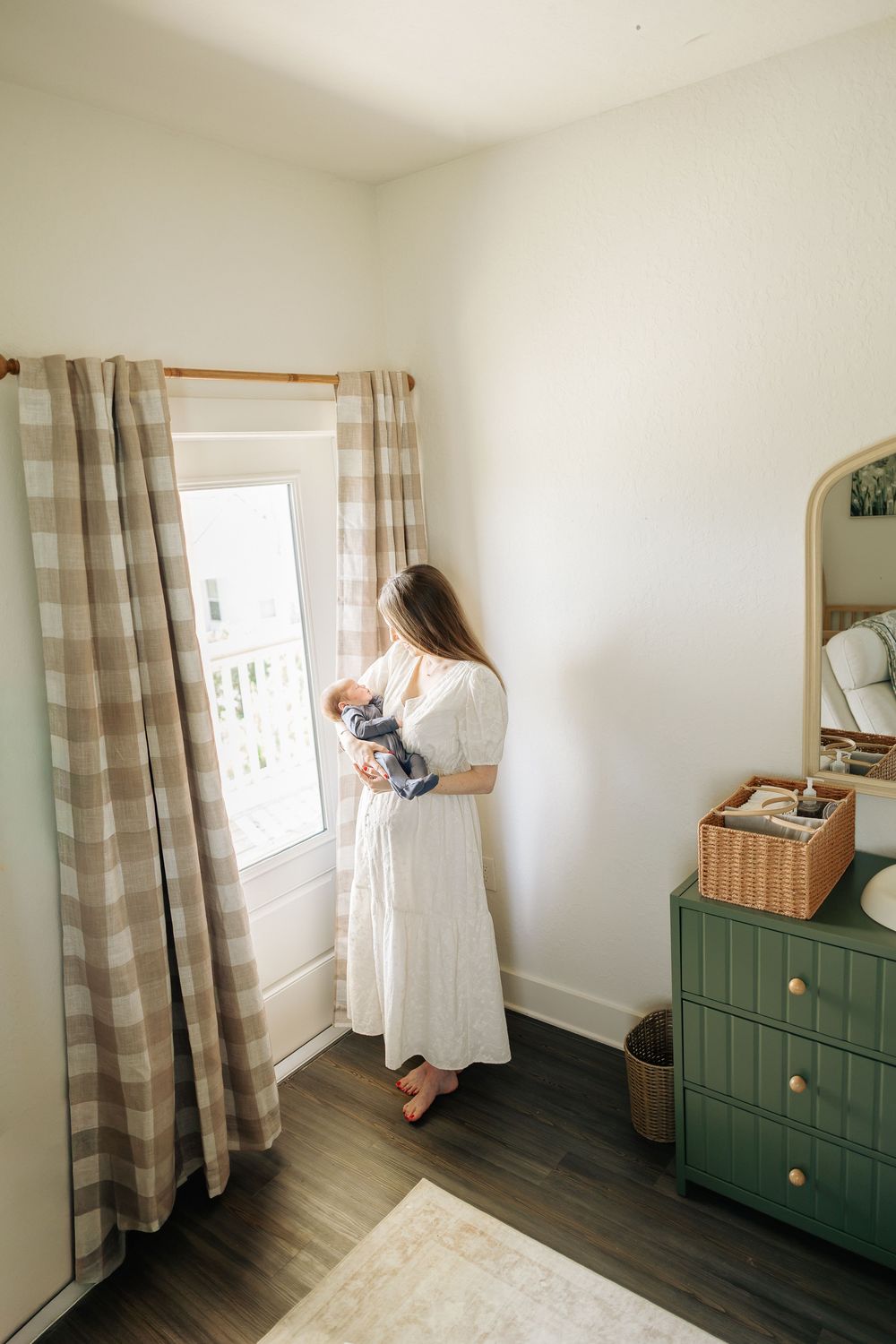 Mother in white dress holds newborn baby by sunlit window in nursery with green dresser and plaid curtains.