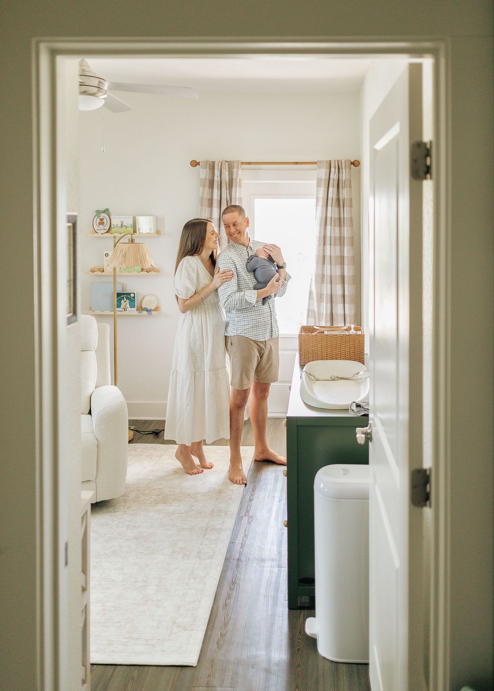 A couple stands in a softly decorated nursery, with the man holding a newborn baby while the woman looks on adoringly.
