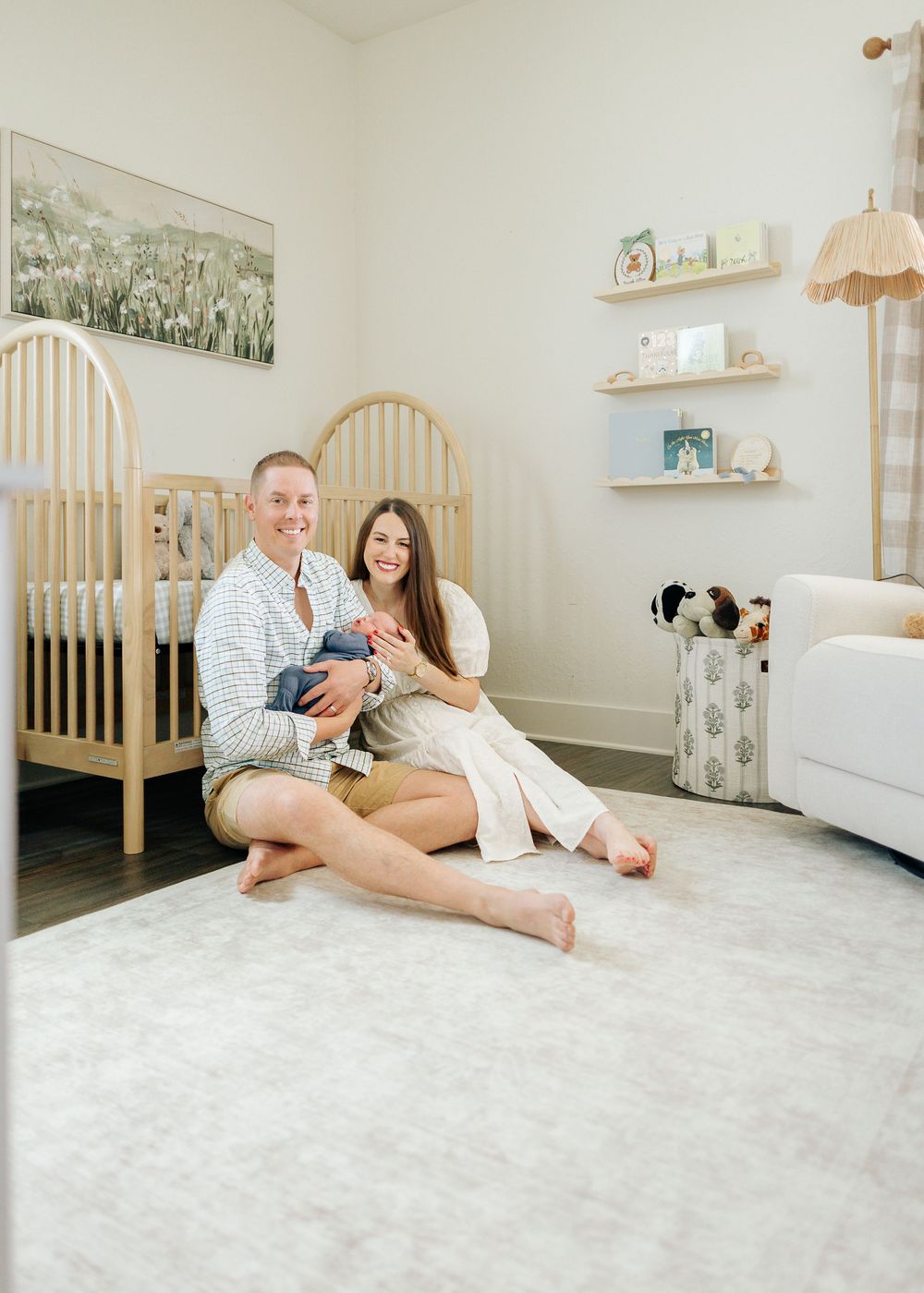 A couple sits on a nursery floor holding their newborn baby, surrounded by a wooden crib, bookshelves, and soft decor.