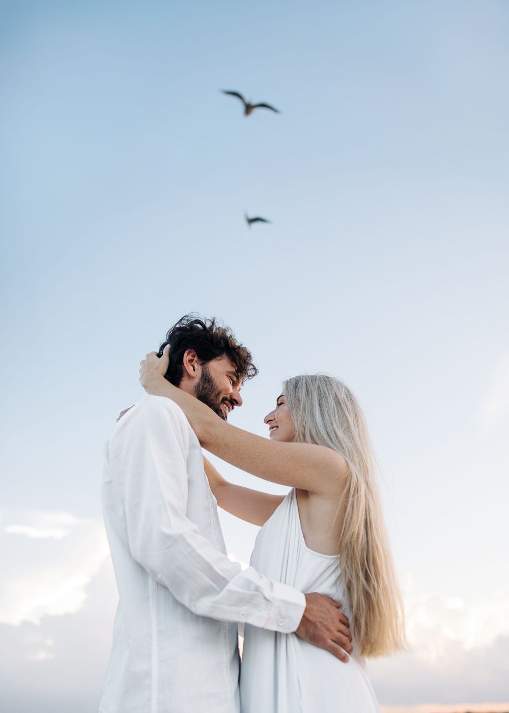 Romantic couple in white embracing on a beach, smiling at each other with birds flying in a blue sky.