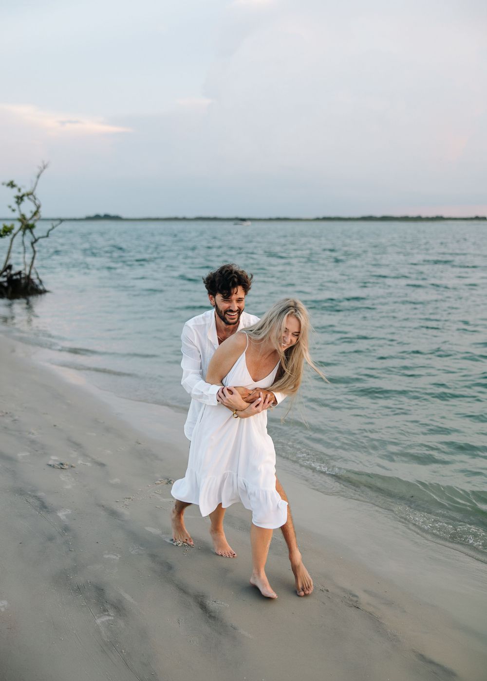 A joyful couple in white outfits walks barefoot along a sandy beach at sunset, laughing and embracing.
