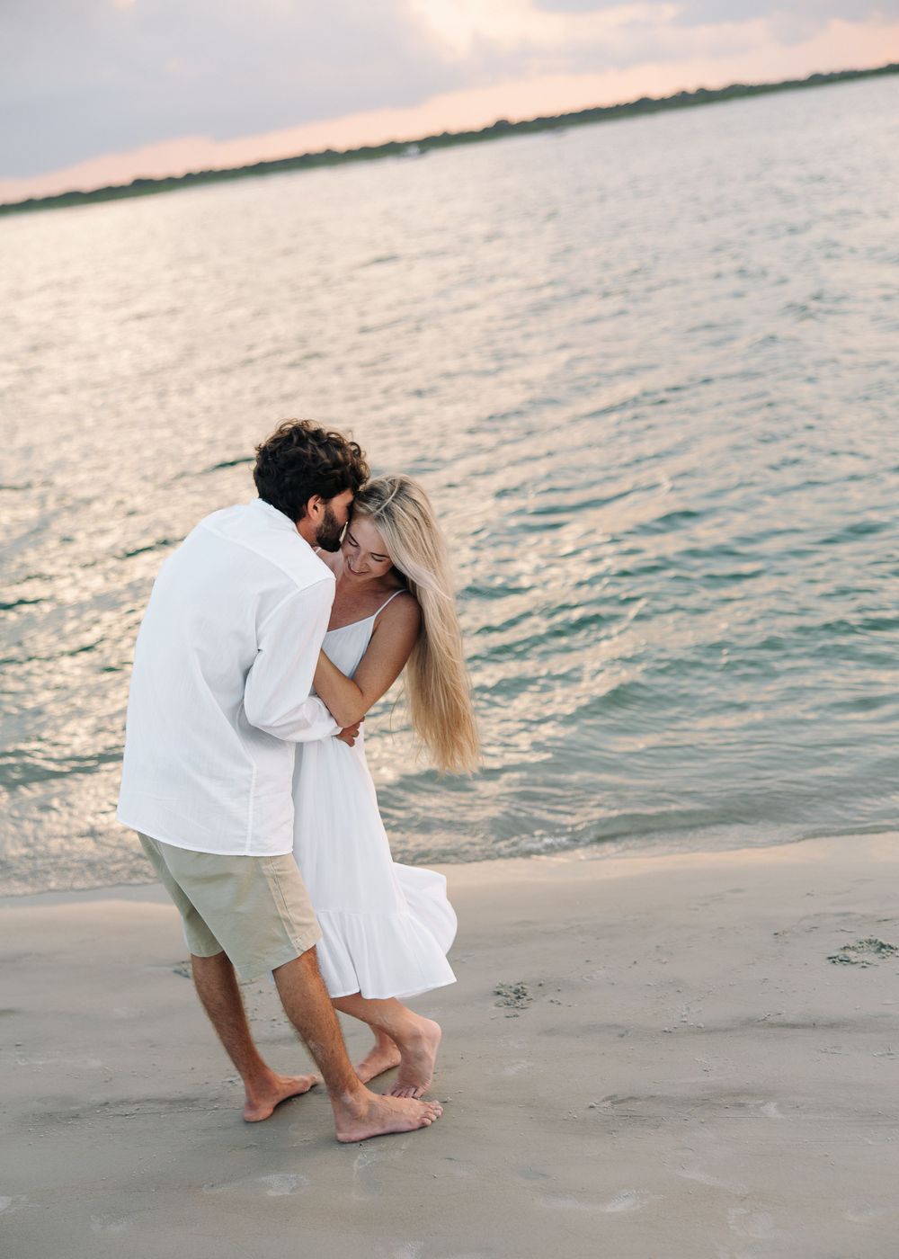 Couple embracing romantically on a sandy beach at sunset, wearing white outfits with ocean waves in the background.