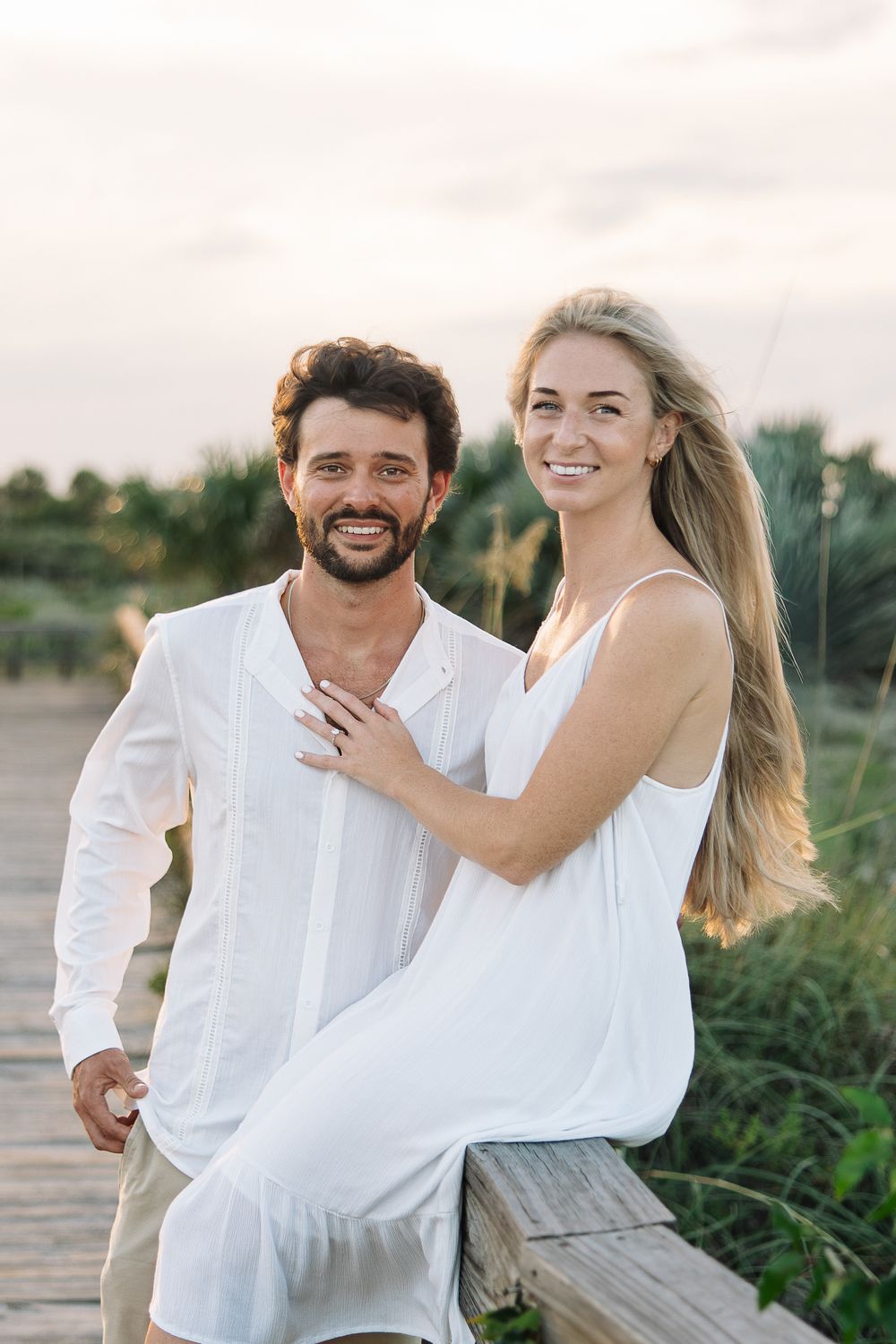 A smiling couple in white outfits embracing on a wooden boardwalk at sunset near lush greenery.