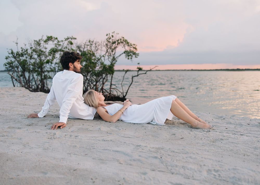 Couple in white outfits relaxing on a sandy beach at sunset, with calm water and mangrove trees in the background.