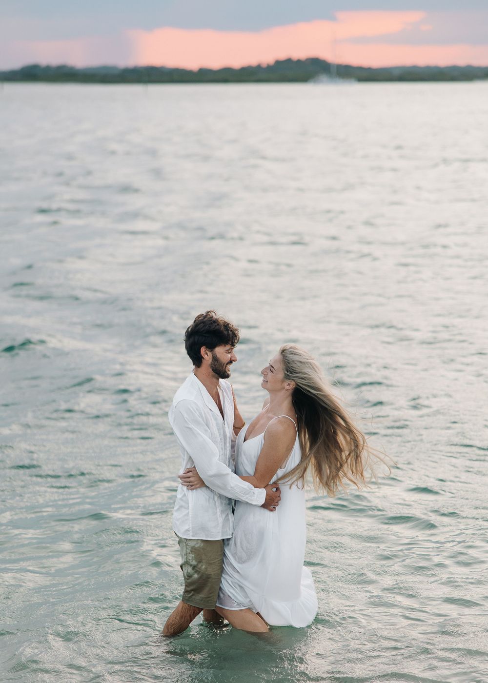 Couple embracing in shallow ocean water at sunset, wearing white outfits, sharing a romantic moment on a tropical beach.