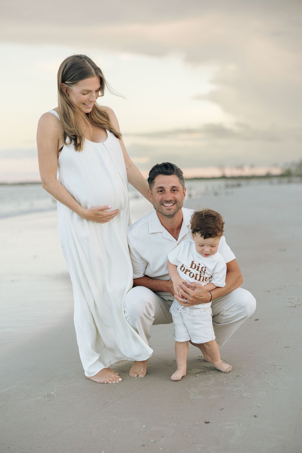 Pregnant woman in white dress with husband and toddler in 'big brother' shirt on beach at sunset.