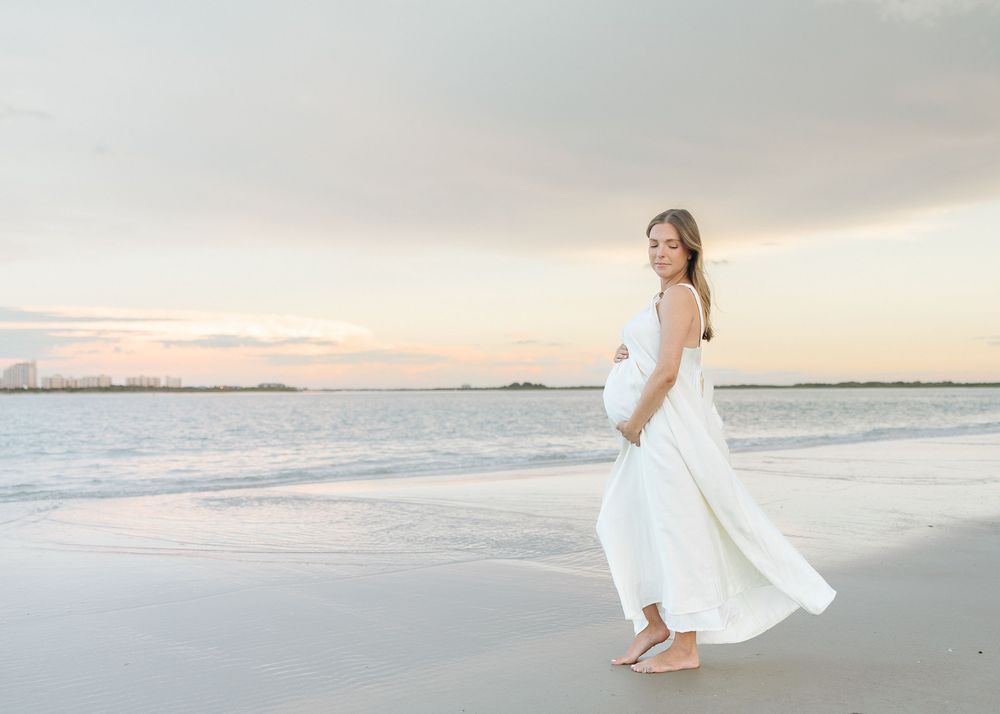 Pregnant woman in white dress on beach at sunset, cradling baby bump with ocean waves in background.