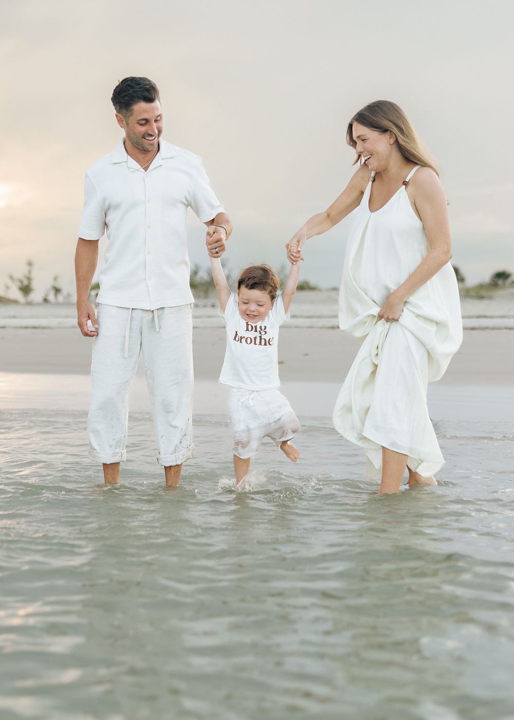 Family of three in white outfits wading in ocean waves at sunset, toddler wearing big brother shirt.
