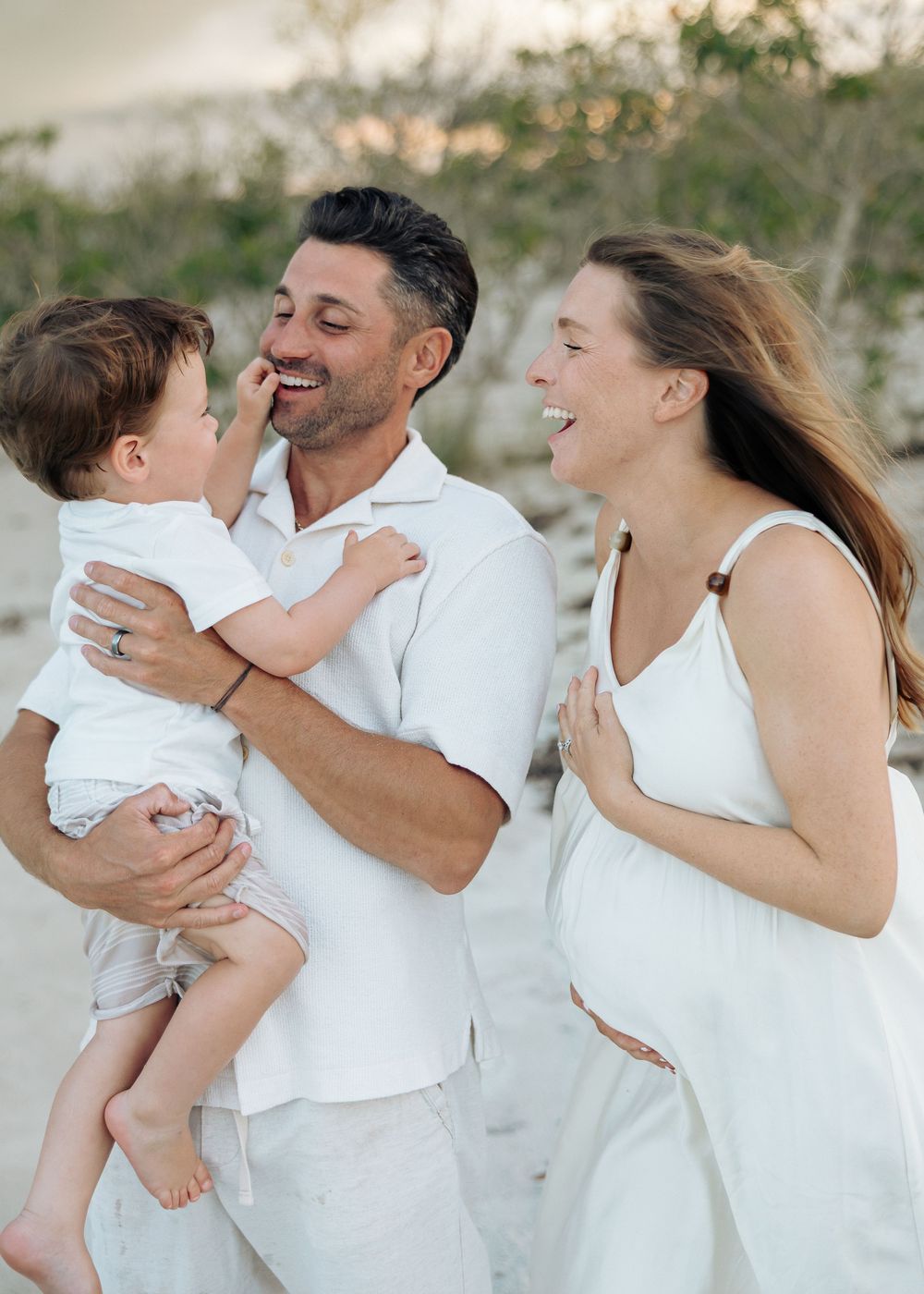 Happy family in white outfits on a beach, parents laughing with their baby boy at sunset.