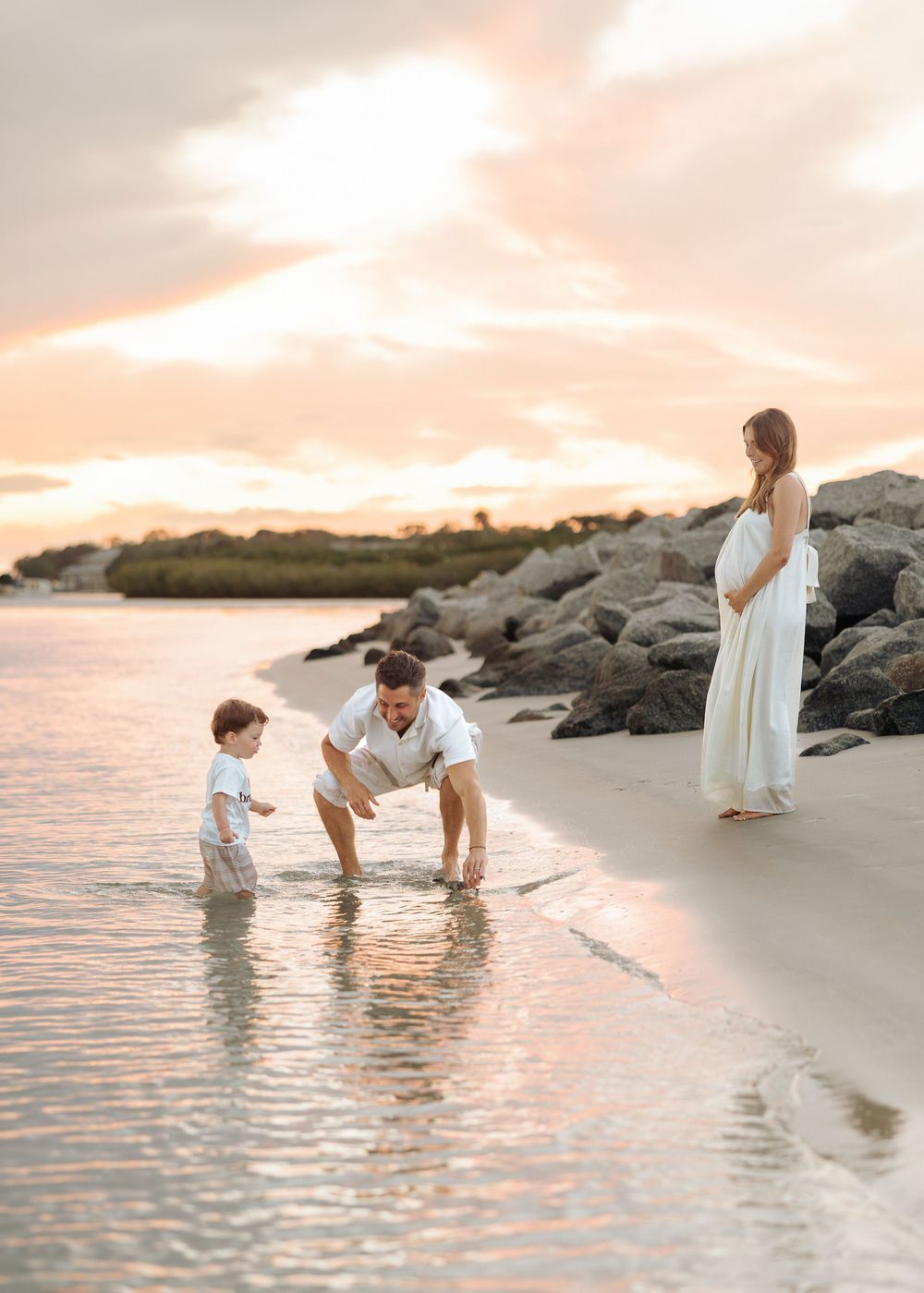 Pregnant woman in white dress watches husband and toddler play in shallow ocean waves at sunset beach.