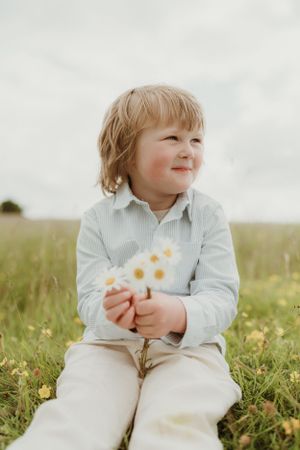 A small child sits in the grass holding wildflowers while wearing light colored clothing.