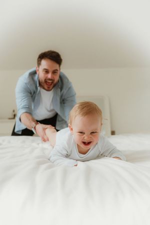 Happy baby crawling on white bedding while adult watches nearby.
