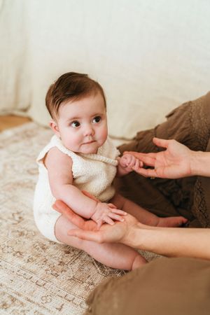 Baby in cream sweater being supported while sitting on textured surface.