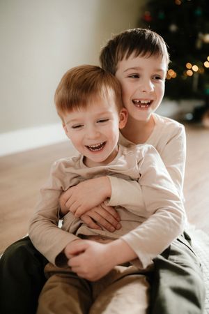 Two young children in white shirts share a joyful embrace while sitting together on a light colored floor.