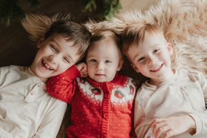 Three children wearing cozy red sweaters share warm hugs and smiles while lying on white bedding.