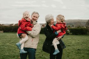Two generations share joy while holding toddlers in matching red coats on a grassy hillside.