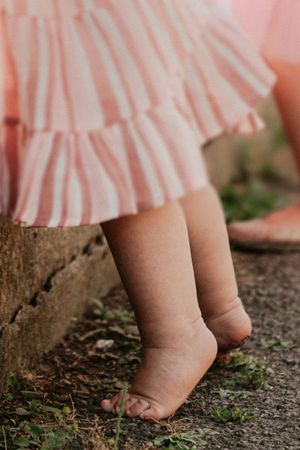 Close-up view of tiny bare baby feet walking on rough concrete ground with pink dress hem visible above.