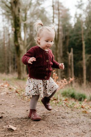A toddler in a burgundy cardigan and floral skirt skips joyfully along a forest trail on an autumn day.