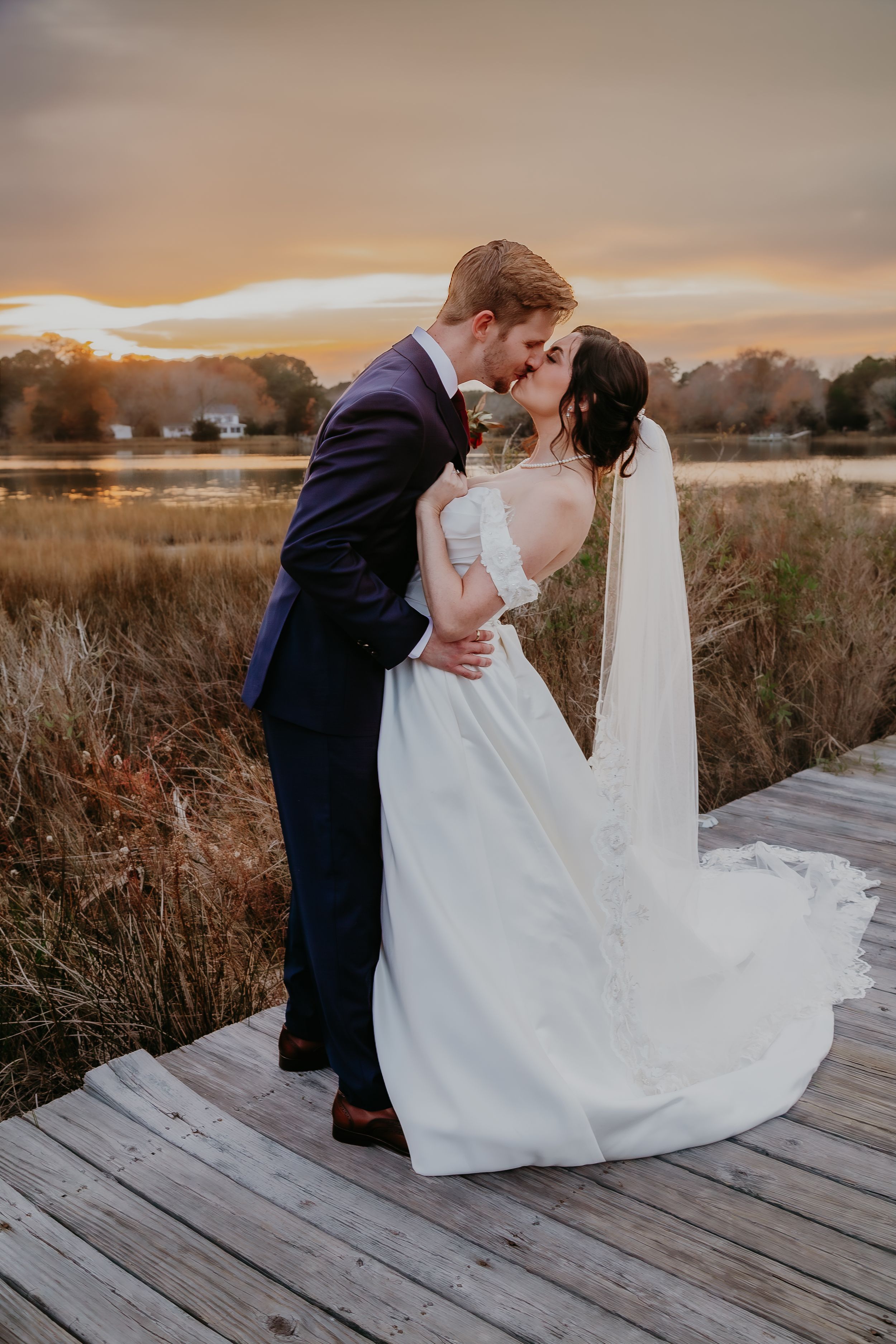 Bride and groom sharing a romantic kiss on wooden dock at golden sunset near a scenic lake.