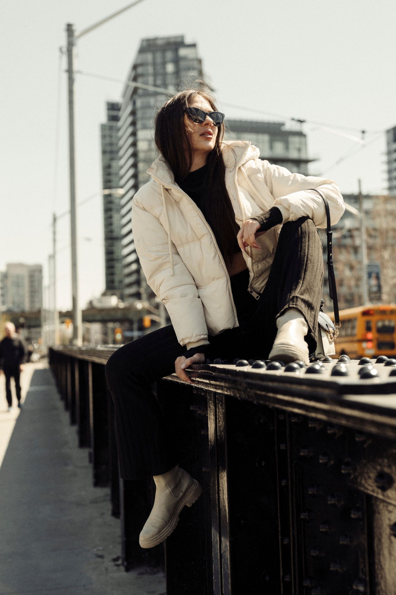 Stylish winter fashion shot with cream puffer jacket and black outfit against urban cityscape with high-rise buildings.